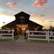 Load image into Gallery viewer, Large wooden barn with string lights at sunset, surrounded by a white fence.
