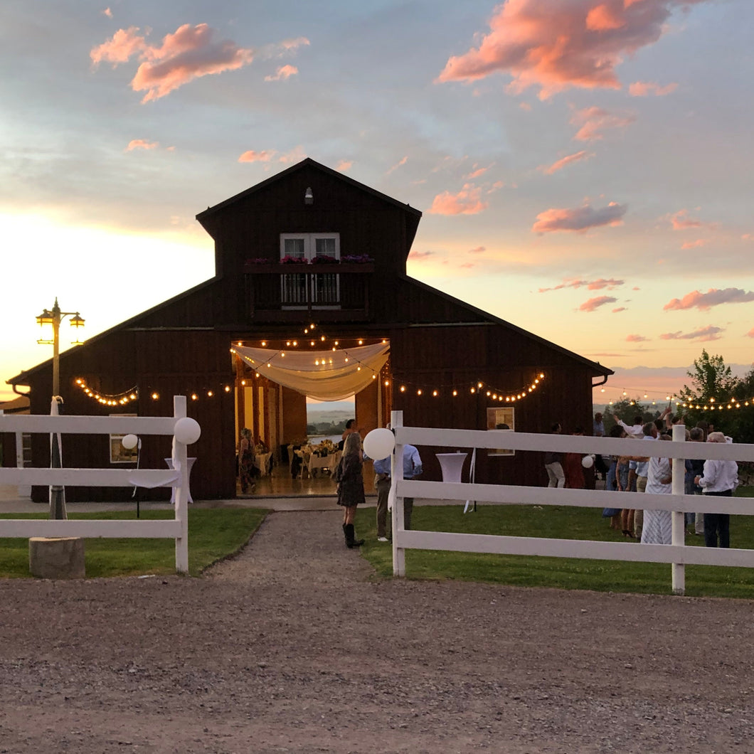 Large wooden barn with string lights at sunset, surrounded by a white fence.