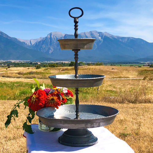 Three-tier metal galvanized serving stand. The round metal trays have tiny gold bead detailing. This sturdy piece has a handle at the top and a sturdy base. The Mission Mountains at the Rugged Horizon wedding venue are in the background.