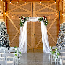 Load image into Gallery viewer, Wedding arch set up indoors for a winter wedding at the Rugged Horizon barn wedding venue in St. Ignatius, Montana. For this wedding, the bride draped chiffon over the arch and added corner bouquets of carnations, white spider mums, and greenery. Frosted Christmas trees with blue and silver ornaments add to the cozy winter look.
