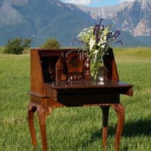 Load image into Gallery viewer, Antique wooden desk with a fold-down top. Store important papers during your wedding ceremony, or tuck your wedding bands into the intricate drawers and cubby holes. Beautiful leaf carvings on the legs. Dark wood stain. The Mission Mountains of the Rugged Horizon wedding venue are in the background.
