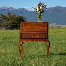 Load image into Gallery viewer, View of the front of the antique wooden podium/desk. It has two drawers and a fold-down top. The legs and base of the desk have intricate wood detailing. Mission Mountains in the background.
